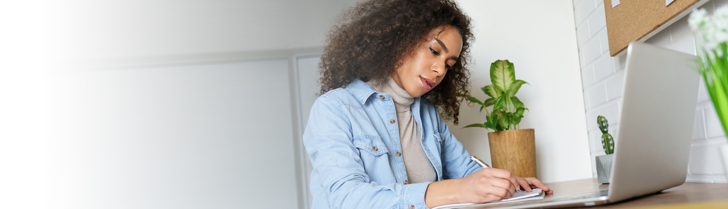 A woman wearing a denim shirt writes in a notebook in front of her laptop.